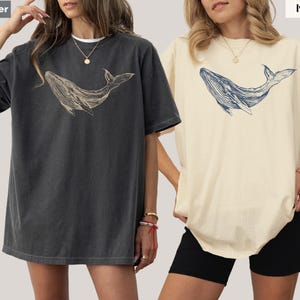 May include: Two women wearing oversized t-shirts with a whale graphic. The shirt on the left is a dark gray color and the shirt on the right is a light ivory color. The whale graphic is in a light beige color on the gray shirt and a dark blue color on the ivory shirt. The text "Pepper" is in the top left corner and the text "Ivory" is in the top right corner.