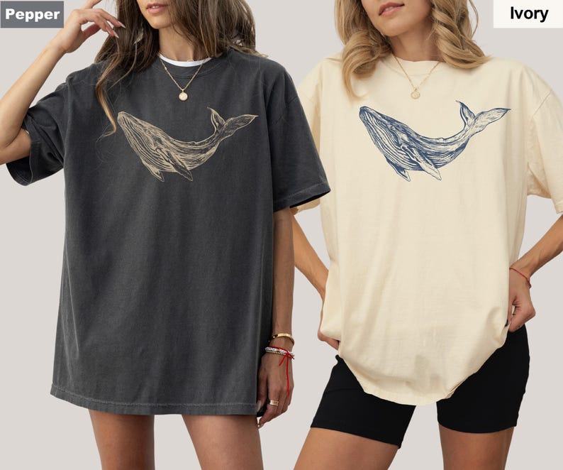 May include: Two women wearing oversized t-shirts with a whale graphic. The shirt on the left is a dark gray color and the shirt on the right is a light ivory color. The whale graphic is in a light beige color on the gray shirt and a dark blue color on the ivory shirt. The text "Pepper" is in the top left corner and the text "Ivory" is in the top right corner.