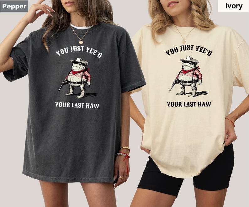Puede incluir: Dos camisetas holgadas, una color pimienta y otra marfil, con un dise&ntilde;o de rana vaquera armada. El texto en las camisetas dice "YOU JUST YEE'D YOUR LAST HAW".