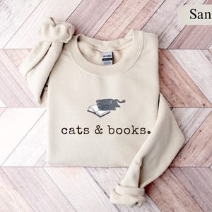 K&ouml;nnte beinhalten: Sandfarbenes Sweatshirt mit Rundhalsausschnitt. Auf der Vorderseite befindet sich eine Grafik einer grauen Katze, die auf einem offenen Buch liegt, mit dem Aufdruck "cats & books" darunter. Die &Auml;rmel sind umgeschlagen.