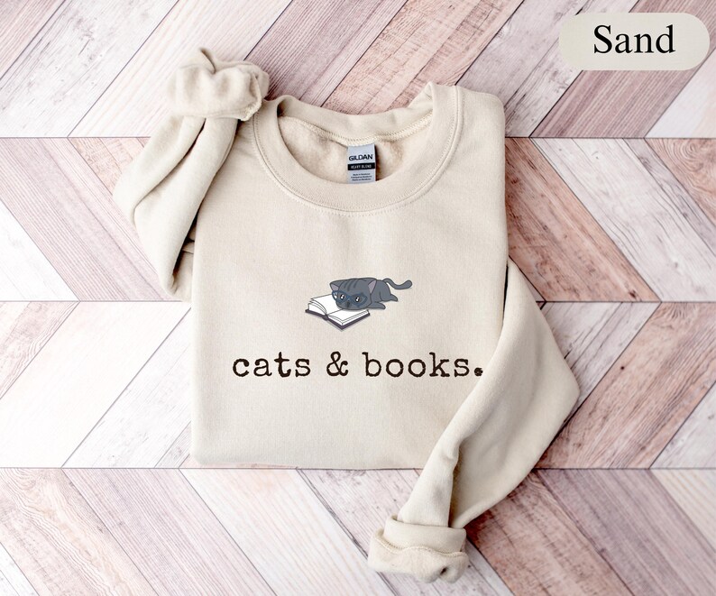 K&ouml;nnte beinhalten: Sandfarbenes Sweatshirt mit Rundhalsausschnitt. Auf der Vorderseite befindet sich eine Grafik einer grauen Katze, die auf einem offenen Buch liegt, mit dem Aufdruck "cats & books" darunter. Die &Auml;rmel sind umgeschlagen.
