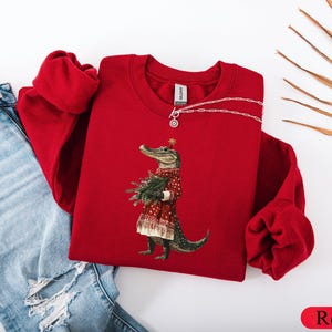 Pu&ograve; includere: Una felpa rossa girocollo con un alligatore che indossa un abito festivo e tiene un albero di Natale. L'alligatore &egrave; il punto focale, con una stella sulla testa. La felpa &egrave; abbinata a jeans blu.