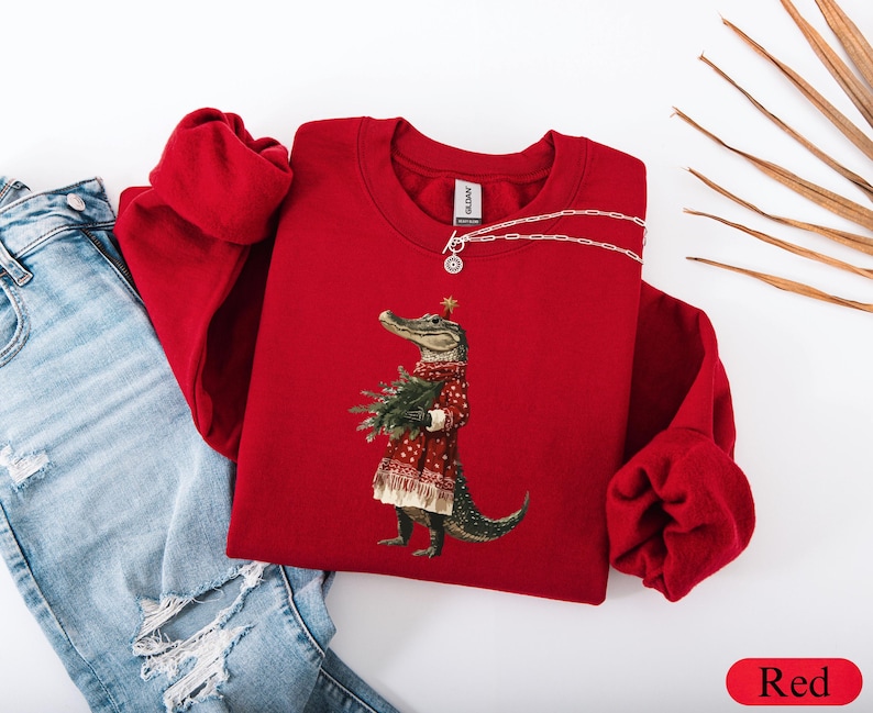 Pu&ograve; includere: Una felpa rossa girocollo con un alligatore che indossa un abito festivo e tiene un albero di Natale. L'alligatore &egrave; il punto focale, con una stella sulla testa. La felpa &egrave; abbinata a jeans blu.