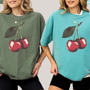 K&ouml;nnte beinhalten: Zwei kurz&auml;rmelige T-Shirts, eines in Moosgr&uuml;n und eines in Meerschaum, jeweils mit einer Grafik von zwei roten Kirschen und einem gr&uuml;nen Blatt. Die Shirts haben eine lockere Passform und einen Rundhalsausschnitt. Die Shirts sind im l&auml;ssigen Stil.