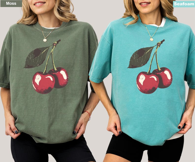 K&ouml;nnte beinhalten: Zwei kurz&auml;rmelige T-Shirts, eines in Moosgr&uuml;n und eines in Meerschaum, jeweils mit einer Grafik von zwei roten Kirschen und einem gr&uuml;nen Blatt. Die Shirts haben eine lockere Passform und einen Rundhalsausschnitt. Die Shirts sind im l&auml;ssigen Stil.