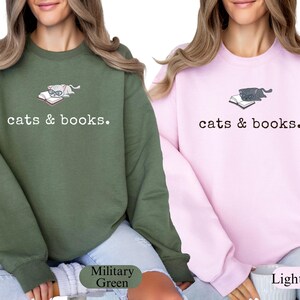 K&ouml;nnte beinhalten: Zwei Sweatshirts, eines in Milit&auml;rgr&uuml;n und eines in Hellrosa, jeweils mit einer Grafik einer Katze mit Brille auf einem Buch. Der Text "cats & books." ist darunter gedruckt. Die Sweatshirts sind lang&auml;rmlig.
