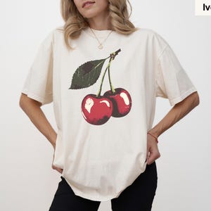 K&ouml;nnte beinhalten: Elfenbeinfarbenes T-Shirt mit einer Grafik von zwei roten Kirschen und einem gr&uuml;nen Blatt. Die Kirschen haben wei&szlig;e Glanzlichter, und das Shirt hat eine lockere Passform. Das Wort "Ivory" ist in einem kleinen Feld oben rechts aufgedruckt.
