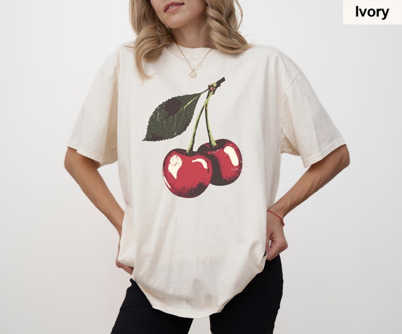 K&ouml;nnte beinhalten: Elfenbeinfarbenes T-Shirt mit einer Grafik von zwei roten Kirschen und einem gr&uuml;nen Blatt. Die Kirschen haben wei&szlig;e Glanzlichter, und das Shirt hat eine lockere Passform. Das Wort "Ivory" ist in einem kleinen Feld oben rechts aufgedruckt.