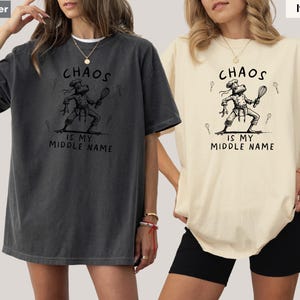 May include: Two t-shirts, one pepper-colored and one ivory-colored, each featuring a graphic of a chef frog holding a whisk. The text "CHAOS IS MY MIDDLE NAME" is printed above and below the frog illustration. The shirts are short-sleeved.