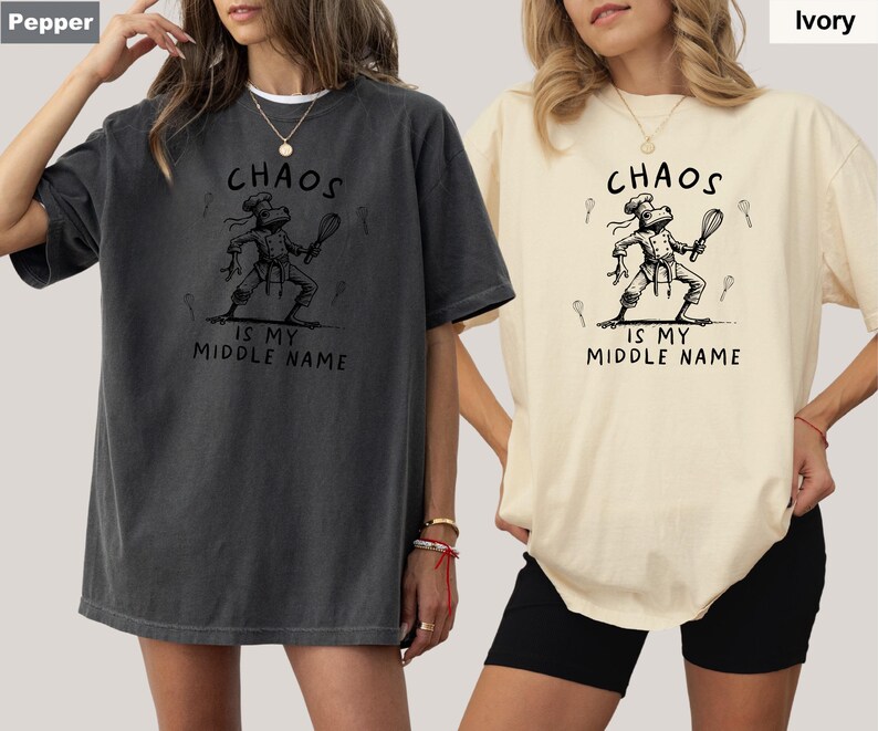 May include: Two t-shirts, one pepper-colored and one ivory-colored, each featuring a graphic of a chef frog holding a whisk. The text "CHAOS IS MY MIDDLE NAME" is printed above and below the frog illustration. The shirts are short-sleeved.