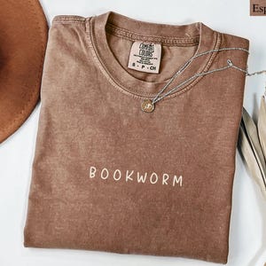 May include: Espresso-colored t-shirt with the word "BOOKWORM" in white lettering. A silver necklace with a small pendant rests on the shirt. The shirt is folded on a white surface, with a brown hat and dried plant visible.