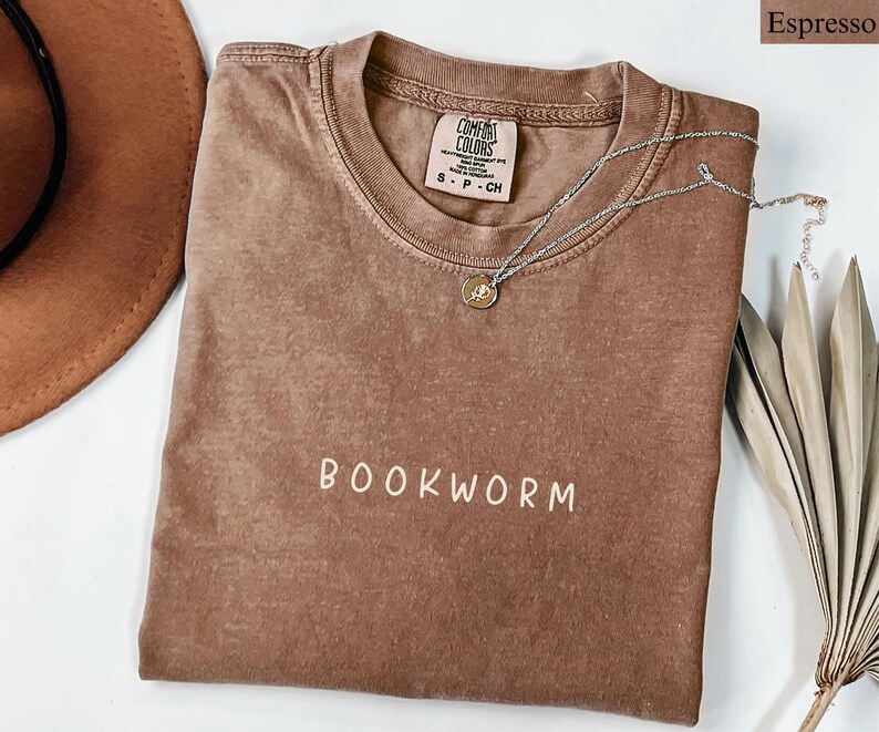 May include: Espresso-colored t-shirt with the word "BOOKWORM" in white lettering. A silver necklace with a small pendant rests on the shirt. The shirt is folded on a white surface, with a brown hat and dried plant visible.