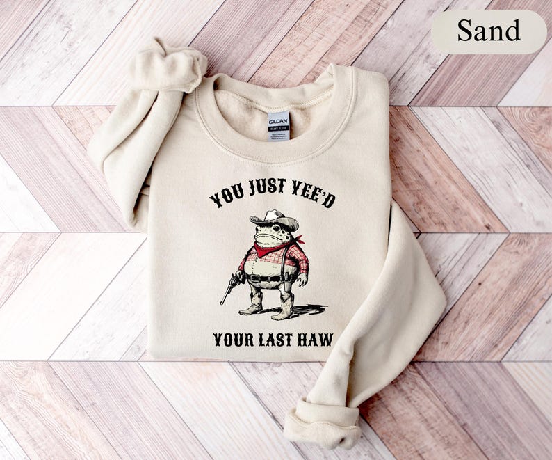Puede incluir: Sudadera de color arena con una rana de dibujos animados vestida de vaquero, sosteniendo una pistola. El texto "YOU JUST YEE'D YOUR LAST HAW" est&aacute; impreso encima y debajo de la ilustraci&oacute;n de la rana. La sudadera tiene cuello redondo y mangas largas.