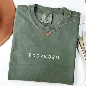 May include: A moss green t-shirt with the word "BOOKWORM" embroidered in white. A gold necklace with a pendant rests on the shirt. A brown hat and dried plant are in the background.