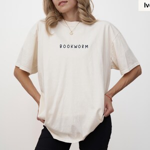 May include: Ivory-colored t-shirt with the word "BOOKWORM" printed in black. The shirt has short sleeves and a relaxed fit. The model is wearing black pants.