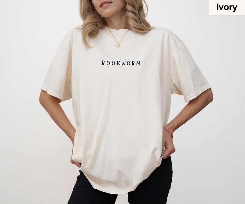 May include: Ivory-colored t-shirt with the word "BOOKWORM" printed in black. The shirt has short sleeves and a relaxed fit. The model is wearing black pants.