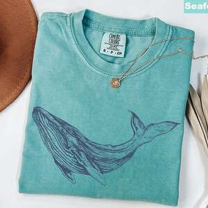 May include: A teal blue t-shirt with a dark blue whale graphic. The shirt has a distressed look and is made of a soft, comfortable fabric. The text "COMFORT COLORS" is printed on a white tag attached to the shirt.