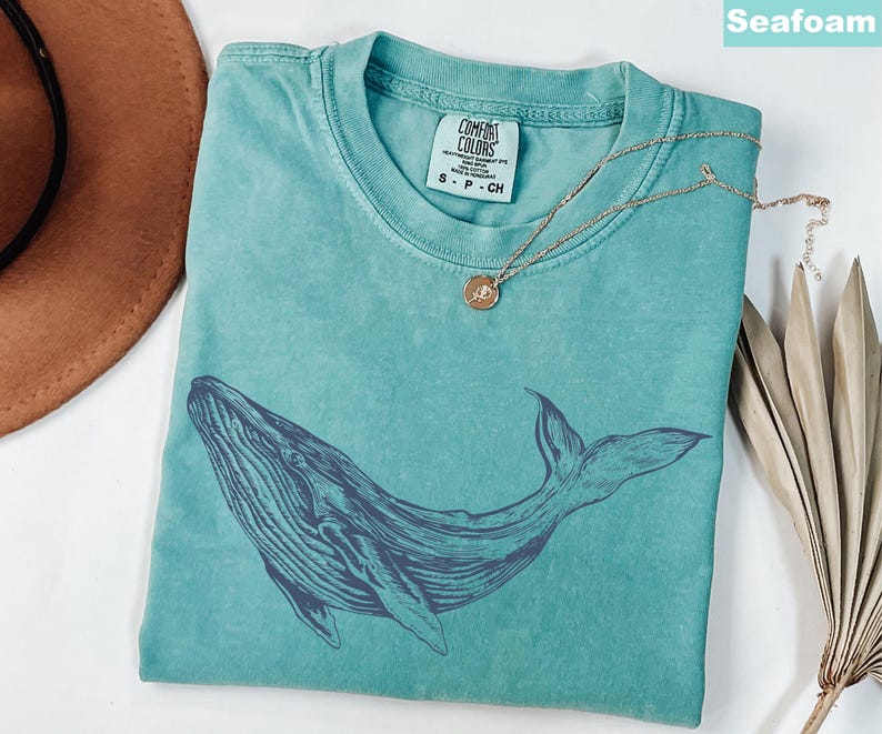 May include: A teal blue t-shirt with a dark blue whale graphic. The shirt has a distressed look and is made of a soft, comfortable fabric. The text "COMFORT COLORS" is printed on a white tag attached to the shirt.