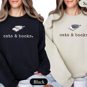 K&ouml;nnte beinhalten: Schwarze und sandfarbene Sweatshirts mit dem Text "cats & books". Jedes Sweatshirt zeigt eine Katzen-Grafik auf einem Buch. Das schwarze Sweatshirt hat eine Katze mit Brille, und das sandfarbene Sweatshirt hat eine graue Katze.