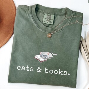 K&ouml;nnte beinhalten: Moosgr&uuml;nes T-Shirt mit der Aufschrift "cats & books". Auf der Vorderseite ist eine Grafik einer Katze mit Brille auf einem Buch abgebildet. Eine goldene Halskette und ein brauner Hut sind ebenfalls im Bild zu sehen.