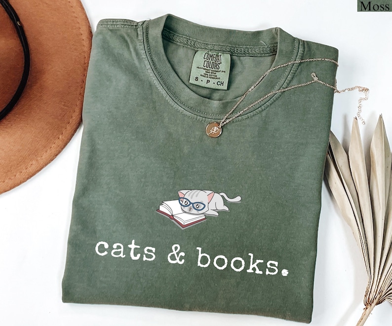 K&ouml;nnte beinhalten: Moosgr&uuml;nes T-Shirt mit der Aufschrift "cats & books". Auf der Vorderseite ist eine Grafik einer Katze mit Brille auf einem Buch abgebildet. Eine goldene Halskette und ein brauner Hut sind ebenfalls im Bild zu sehen.
