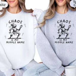 May include: White and sport grey sweatshirts with the text "Chaos is my middle name." The graphic features a frog chef holding a whisk and wearing a chef's hat. The sweatshirts are a crew neck style.
