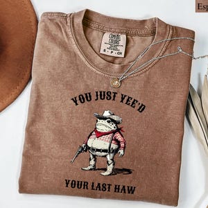 Puede incluir: Camiseta color espresso con un gr&aacute;fico de una rana con sombrero y chaleco de vaquero sosteniendo una pistola. El texto "YOU JUST YEE'D YOUR LAST HAW" est&aacute; impreso encima y debajo de la ilustraci&oacute;n de la rana. Un collar plateado est&aacute; sobre la camiseta.