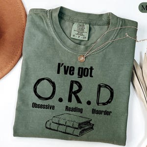 May include: A moss green t-shirt with the text "I've got O.R.D. Obsessive Reading Disorder" in black. The shirt has a crew neck and short sleeves. A gold necklace with a pendant is draped across the shirt.