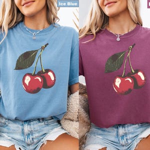 K&ouml;nnte beinhalten: Zwei kurz&auml;rmelige T-Shirts, eines in Eisblau und eines in Beere, jeweils mit einer Grafik von zwei roten Kirschen mit gr&uuml;nen Bl&auml;ttern. Die Shirts sind aus weichem Material und haben eine lockere Passform, geeignet f&uuml;r Freizeitkleidung.