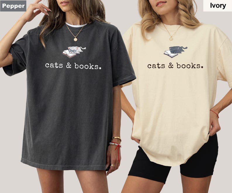K&ouml;nnte beinhalten: Zwei &uuml;bergro&szlig;e T-Shirts, eines in Pfeffer und eines in Elfenbein, mit einer Grafik einer Katze auf einem Buch und dem Text "cats & books". Die Shirts haben kurze &Auml;rmel und eine lockere Passform.