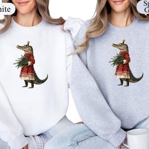 Pu&ograve; includere: Felpe bianche e grigio sportivo con una stravagante illustrazione di un alligatore vestito con un abito festivo rosso, che tiene un piccolo albero di Natale. L'alligatore indossa un cappello festivo. Le felpe sono a girocollo.