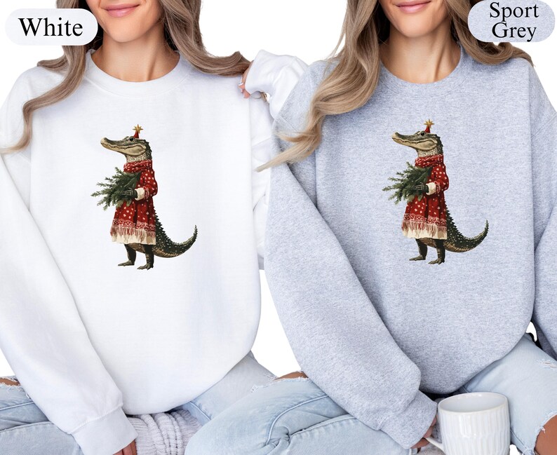 Pu&ograve; includere: Felpe bianche e grigio sportivo con una stravagante illustrazione di un alligatore vestito con un abito festivo rosso, che tiene un piccolo albero di Natale. L'alligatore indossa un cappello festivo. Le felpe sono a girocollo.