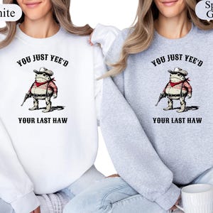 Puede incluir: Sudaderas blancas y gris deportivo con un gr&aacute;fico de rana vaquera de dibujos animados. La rana lleva sombrero, pa&ntilde;uelo rojo y sostiene una pistola. El texto en las sudaderas dice "YOU JUST YEE'D" y "YOUR LAST HAW".