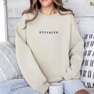 May include: Sand-colored sweatshirt with the word "BOOKWORM" printed in black. The sweatshirt is a neutral color and has a crew neck. The person in the image is wearing blue jeans and is holding a white mug.