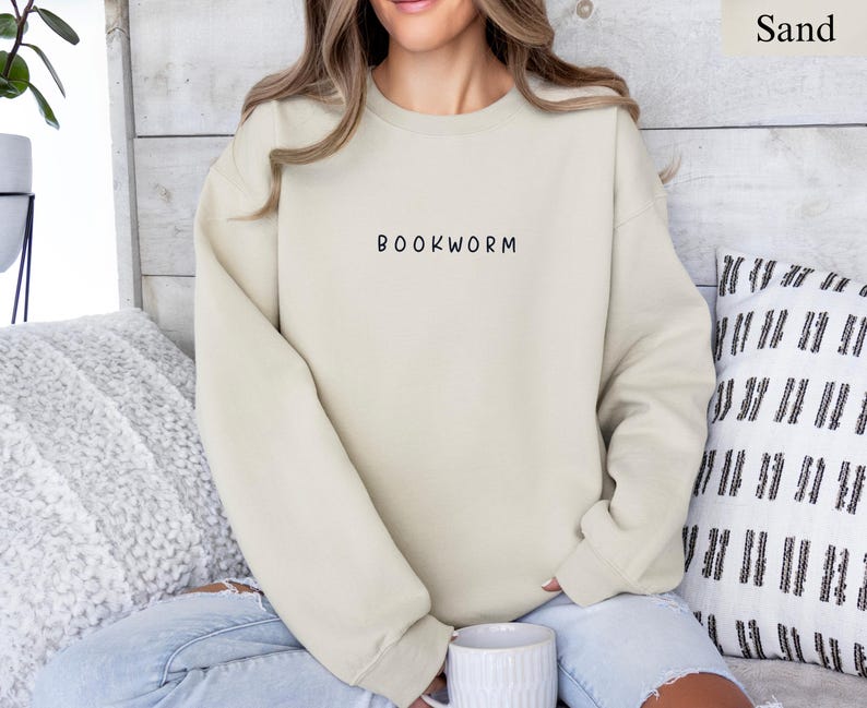 May include: Sand-colored sweatshirt with the word "BOOKWORM" printed in black. The sweatshirt is a neutral color and has a crew neck. The person in the image is wearing blue jeans and is holding a white mug.