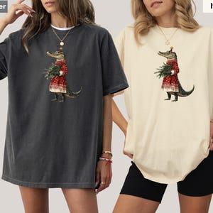 Pu&ograve; includere: Due magliette oversize, una color pepe e una avorio, con un'illustrazione stravagante. La grafica raffigura un alligatore in un vestito rosso che tiene del verde. L'alligatore indossa un cappello festivo, suggerendo un tema festivo.