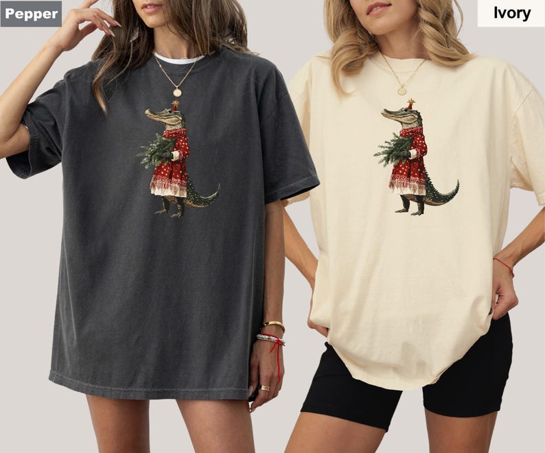 Pu&ograve; includere: Due magliette oversize, una color pepe e una avorio, con un'illustrazione stravagante. La grafica raffigura un alligatore in un vestito rosso che tiene del verde. L'alligatore indossa un cappello festivo, suggerendo un tema festivo.