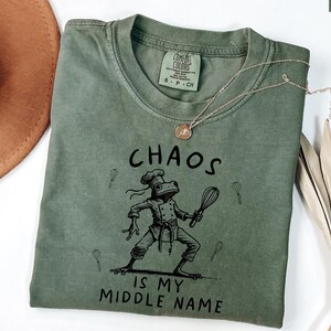 May include: Moss green t-shirt with the text "CHAOS IS MY MIDDLE NAME" and a cartoon frog chef holding a whisk. The shirt has a crew neck and short sleeves. A gold necklace is draped across the shirt.