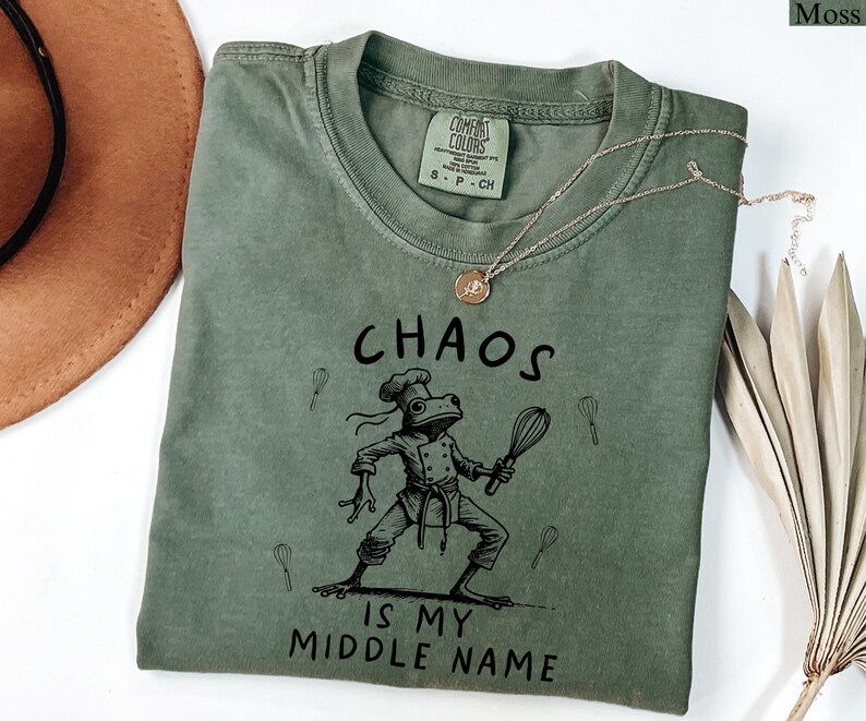 May include: Moss green t-shirt with the text "CHAOS IS MY MIDDLE NAME" and a cartoon frog chef holding a whisk. The shirt has a crew neck and short sleeves. A gold necklace is draped across the shirt.