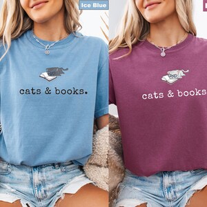 K&ouml;nnte beinhalten: Zwei kurz&auml;rmelige T-Shirts, eines in Eisblau und eines in Beere, jeweils mit dem Schriftzug "cats & books". Die Shirts zeigen eine Grafik einer Katze, die auf einem Buch liegt. Das eisblaue Shirt hat eine dunkelgraue Katze, w&auml;hrend das Beerenshirt eine Brille tragende Katze hat.