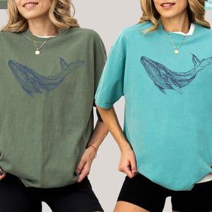 May include: Two women wearing oversized t-shirts with a blue whale graphic. The t-shirt on the left is moss green and the t-shirt on the right is seafoam blue.