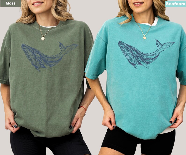May include: Two women wearing oversized t-shirts with a blue whale graphic. The t-shirt on the left is moss green and the t-shirt on the right is seafoam blue.