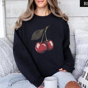 K&ouml;nnte beinhalten: Schwarzes Sweatshirt mit einer Grafik von zwei roten Kirschen und einem gr&uuml;nen Blatt. Die Kirschen haben einen Vintage-Look mit leichtem Used-Effekt. Das Sweatshirt ist einfarbig schwarz.