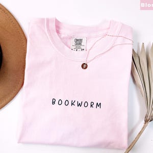 May include: A light pink t-shirt with the word "BOOKWORM" printed in black. A gold necklace with a circular pendant rests on the shirt. A brown hat and dried palm leaf are in the background.