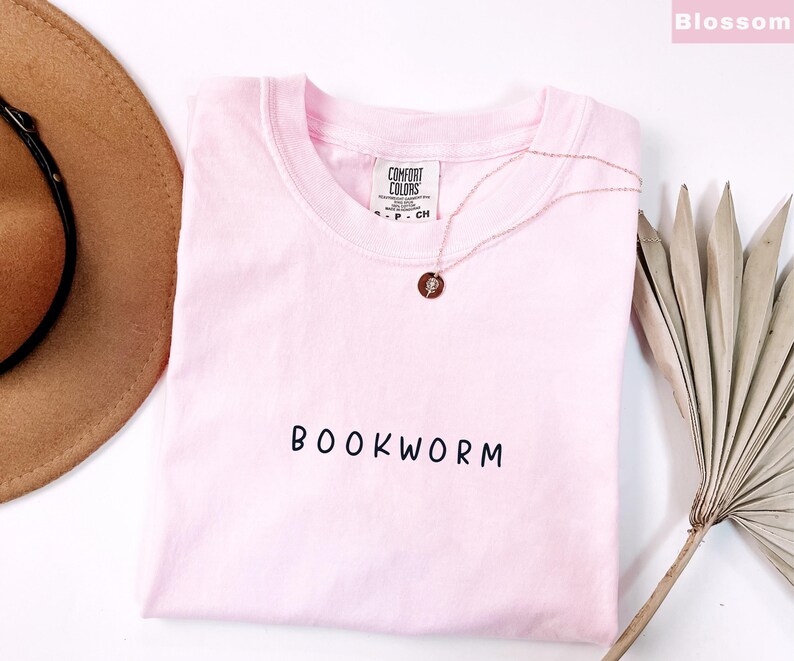 May include: A light pink t-shirt with the word "BOOKWORM" printed in black. A gold necklace with a circular pendant rests on the shirt. A brown hat and dried palm leaf are in the background.