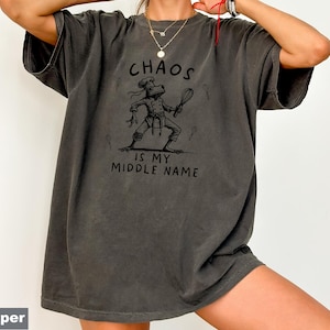 May include: Dark gray t-shirt with the text "CHAOS IS MY MIDDLE NAME" and a cartoon frog chef graphic. The frog is wearing a chef's hat and holding a whisk. The shirt is a relaxed fit.