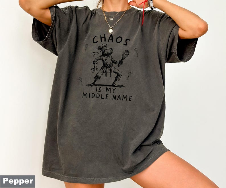 May include: Dark gray t-shirt with the text "CHAOS IS MY MIDDLE NAME" and a cartoon frog chef graphic. The frog is wearing a chef's hat and holding a whisk. The shirt is a relaxed fit.