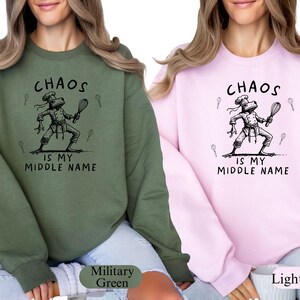 May include: Two sweatshirts, one military green and one light pink, each featuring a black graphic of a frog chef holding a whisk. The text "CHAOS IS MY MIDDLE NAME" is above and below the frog illustration. The sweatshirts are casual wear.