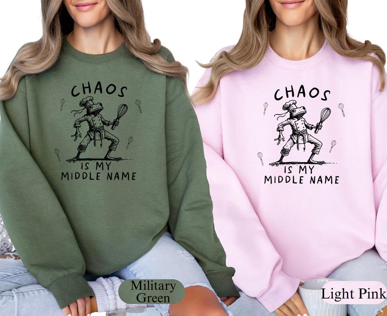 May include: Two sweatshirts, one military green and one light pink, each featuring a black graphic of a frog chef holding a whisk. The text "CHAOS IS MY MIDDLE NAME" is above and below the frog illustration. The sweatshirts are casual wear.