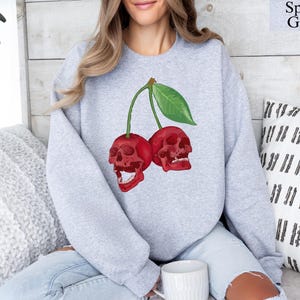 Może przedstawiać: Jasnoszara bluza Sport Grey z efektownym wzorem dw&oacute;ch czerwonych wiśni w kształcie czaszki i zielonych liści. Grafika znajduje się z przodu. Tekst "Sport Grey" znajduje się w prawym g&oacute;rnym rogu.